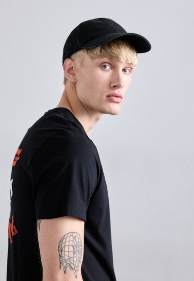 Jeune homme blond portant une casquette noire et un t-shirt noir regarde par-dessus son épaule, montrant un tatouage de globe en train de fondre sur son bras supérieur, fond gris.