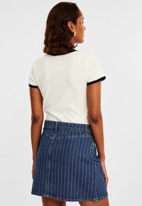 Weißes geripptes T-Shirt mit schwarzem Saum, kombiniert mit einem blauen Denim-Rock mit vertikalen weißen Streifen und einem Bund. Rückansicht dargestellt.