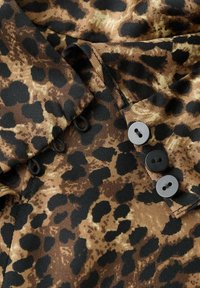 Material cu imprimeu de leopard în nuanțe de maro și negru, cu guler și trei nasturi negri în față pentru închidere.