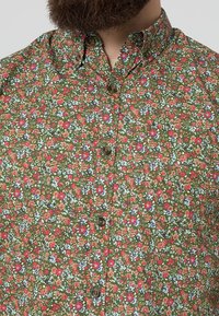 Groene button-up shirt met een bloemmotief, in combinatie met kleine rode, blauwe en witte bloemen, en bruine knopen aan de voorkant.