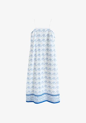 Une femme se tient contre un mur, portant une robe blanche avec des motifs de vagues bleues et des noms d'îles grecques, des sandales et des créoles dorées.