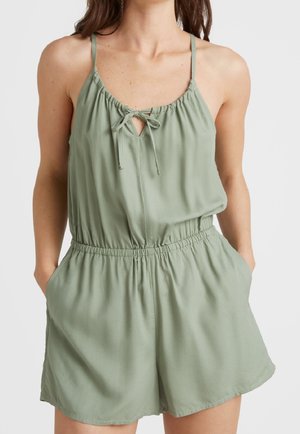 Nainen pukeutuneena vaaleanvihreään, hihattomaan haalariin, jossa on joustava vyötärö, ohuet olkaimet, edessä solmimisnauha ja taskut, seisoo käsin taskuissa.