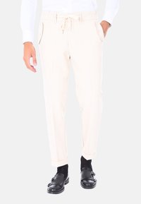 Pantaloni beige con vita regolabile, tasche laterali e orli cuffiti. Il tessuto appare liscio e leggero. Indossati con scarpe nere e calzini.
