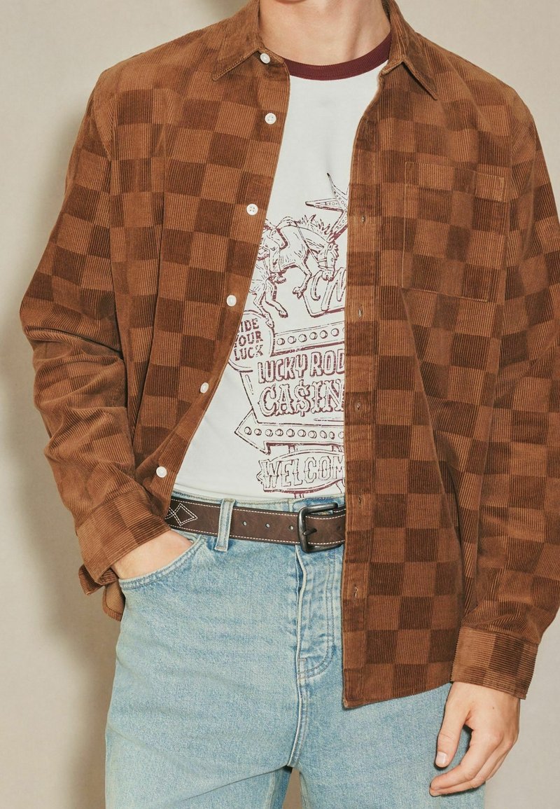 Homme portant un jean bleu clair, une ceinture marron, un t-shirt blanc avec un motif graphique, et une chemise marron à carreaux ouverte, main dans la poche.