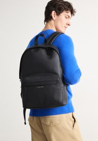 Zaino in pelle nera con una texture liscia, forma arrotondata, tasca frontale con zip e logo Calvin Klein. Straps regolabili e manico superiore visibili.