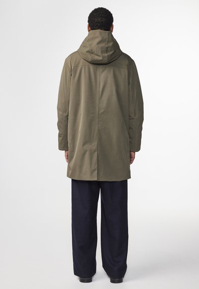 Olivgrön vattentät parkas med huva, rak skärning och långa ärmar. Bärs över mörka randiga byxor, visar minimala designdetaljer.