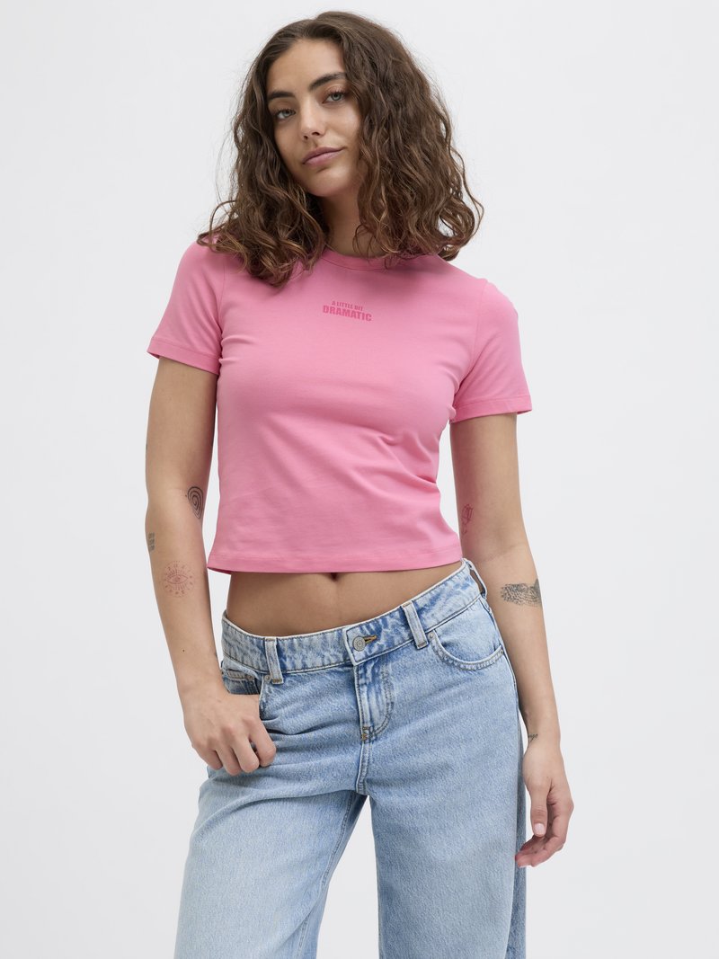 Tettsittende, kortermet, rosa crop-top med teksten "A LITTLE BIT DRAMATIC". Kombinert med lyseblå, avslappede jeans. Glatt tekstur.