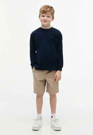 Ragazzo in piedi con le mani nelle tasche, indossa una maglia blu navy a maniche lunghe, pantaloncini beige, calzini bianchi e sneakers bianche, sorridendo leggermente.