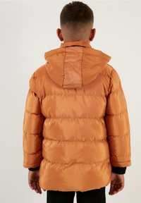 Chaqueta acolchada en color naranja, con capucha, textura acolchada y puños negros. La parte trasera presenta un acabado liso y brillante con detalles mínimos.