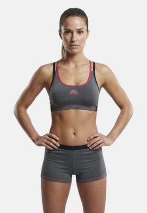 Donna in forma con le mani sui fianchi, indossa un reggiseno sportivo grigio e pantaloncini abbinati con rifiniture rosa, su uno sfondo bianco semplice.