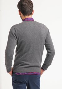 Grauer Strickpullover mit einem runden Halsausschnitt und gerippten Bündchen, über einem karierten Hemd mit roten und blauen Tönen getragen. Einfaches und sauberes Design.