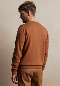 Brązowy sweter dzianinowy z okrągłym dekoltem, żebrowanymi mankietami i dołem. Noszony z jasnobeżowymi spodniami, prezentującymi gładką fakturę i dopasowany krój.