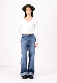 Bela rebrasta dolgi rokav top s krojenim ovratnikom v obliki V, v kombinaciji z modrimi flare jeans hlačami z nedokončanim robom, obut z črnimi platform čevlji.