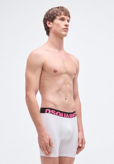 Calzoncillos bóxer blancos con una cintura negra que presenta un texto en rosa intenso "DSQUARED". El material parece suave con una textura lisa.
