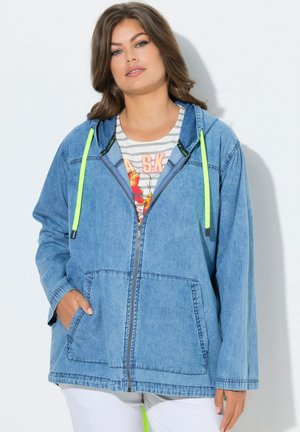 Kobieta nosząca jasnoniebieską dzianinową bluzę z kapturem na suwak i neonowozielonymi sznurkami oraz pasiastą kwiatową koszulę pod spodem.