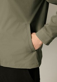 Olivenfarvet letvægtsjakke med en glat tekstur, der har en sidelomme og en skræddersyet ærmekant. Enkel, ren design.