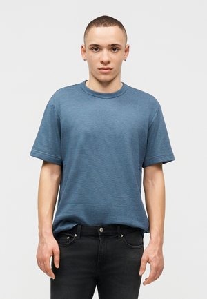 Young person with short hair wearing a blue striped t-shirt and black jeans, standing against a plain light background.