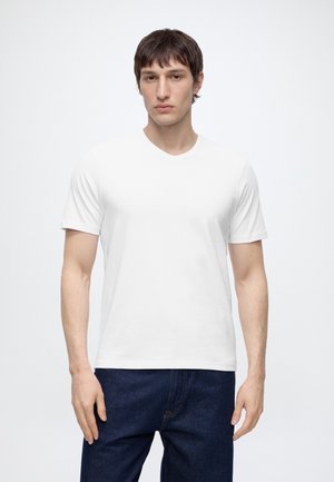 Jeune homme aux cheveux bruns courts portant un t-shirt blanc uni à manches courtes et un jean bleu foncé, debout devant un fond blanc.
