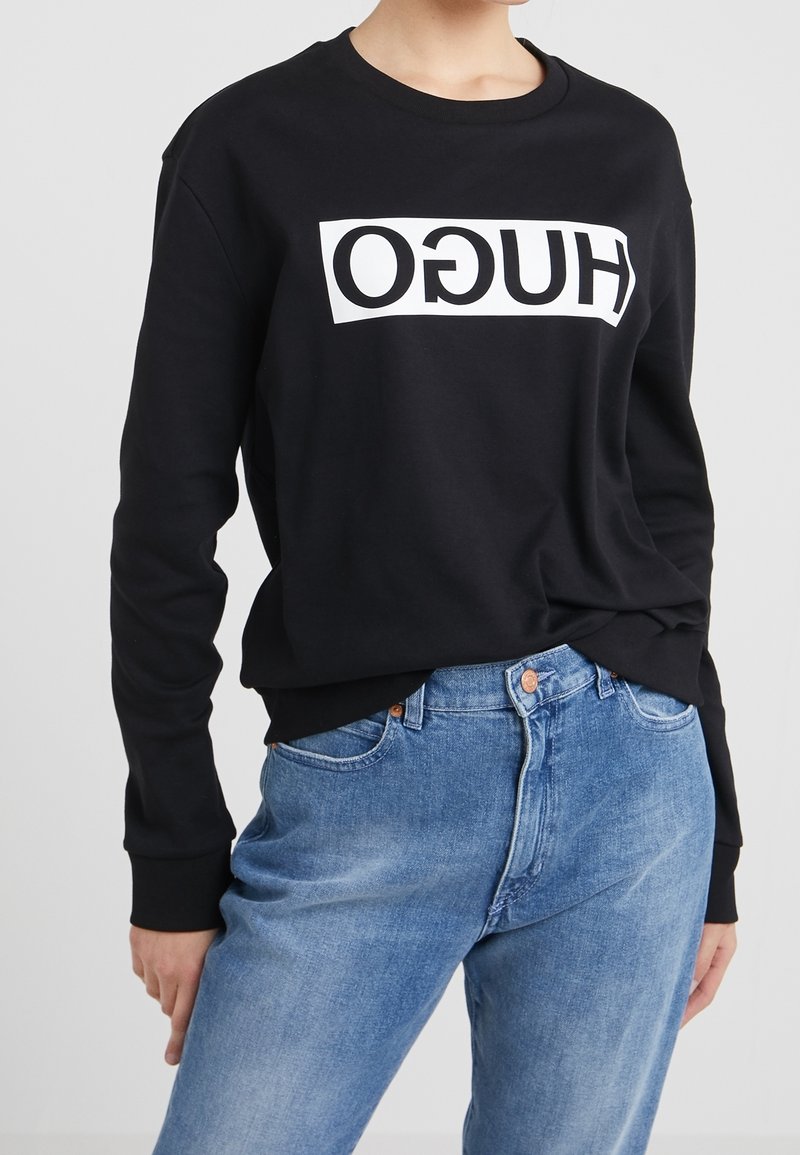 Persona que lleva un sudadera negra con el logotipo en blanco "HUGO" al revés y unos vaqueros de denim azul, de pie frente a un fondo simple.