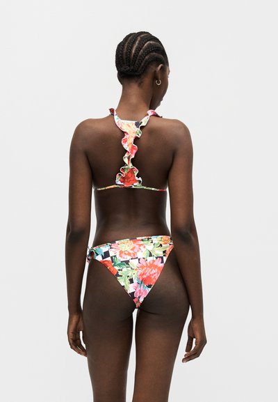 Γυναίκα με πλεξούδες στα μαλλιά, που φοράει μπικίνι με λουλουδάτα βολάν, στέκεται γυρισμένη με την πλάτη σε ένα μονόχρωμο γκρι φόντο.