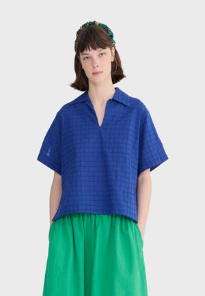 Frau trägt ein blaues kariertes Hemd mit kurzen Ärmeln und Kragen sowie einen leuchtend grünen Rock, Hände in den Taschen und ein dunkles Blumen-Haarband.