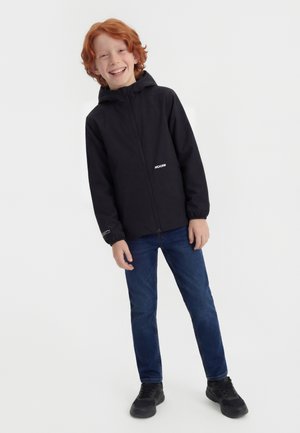 Naeratav punapea poiss, kes kannab musta kapuutsiga jopet, siniseid teksapükse ja musti tossusid, seisab valgel taustal.