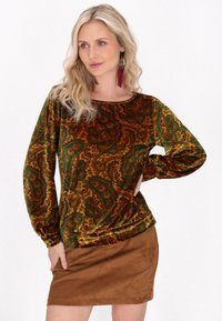 Grüne und goldene Paisley-Print-Velours langärmelige Bluse kombiniert mit einem braunen Wildleder-Minirock. Auffällige Texturen und filigrane Muster sind vorhanden.