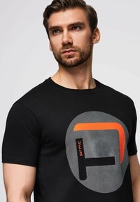 Černé tričko s krátkým rukávem s kruhovým grafickým designem a tečkovanými vzory, vynikající oranžovým akcentem. Měkká bavlněná látka.