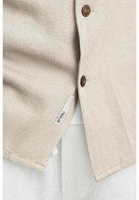 Beige overhemd met knopen in bruin, met een gestructureerde stof en een label aan de zijkant. Gelaagd met een witte binnenhemd dat eronder zichtbaar is.