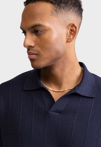 Νεαρός με κοντά σγουρά μαλλιά που φοράει ένα υφασμάτινο μπλε πουκάμισο με γιακά και λεπτή χρυσή αλυσίδα στο λαιμό, κοιτάζοντας προς τα αριστερά.