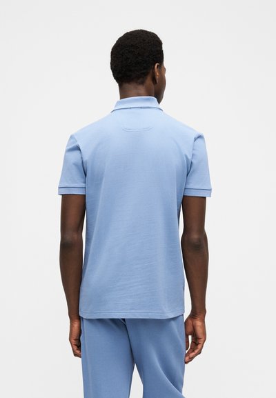 Homme aux cheveux courts et bouclés portant un polo bleu clair à manches courtes et un pantalon assorti, vu de dos sur un fond uni.