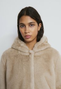 Abrigo de piel sintética beige con capucha, que presenta una textura suave y un cierre frontal. Diseño sencillo con escote redondeado.