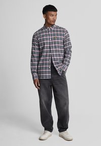 Camicia button-up di colore verde scuro con motivo a quadri in rosso, blu e bianco. Presenta un colletto, maniche lunghe e un logo sul petto sinistro.