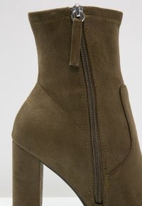 Bottine en daim vert olive à talon haut, dotée d'une fermeture éclair latérale et d'un design minimaliste, mettant en valeur une texture lisse et des détails cousus.