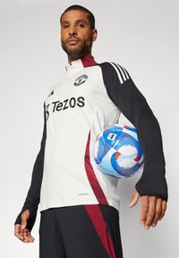 White sports top with black sleeves and maroon accents, featuring a collar, logo, and "Tezos" text. Player holds a blue and white patterned soccer ball.