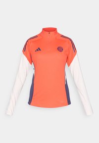 Hosszú ujjú sportfelső, narancssárga testtel, fehér ujjal, lila díszítésekkel, fél cipzáras gallérral és logóval a mellrészen.