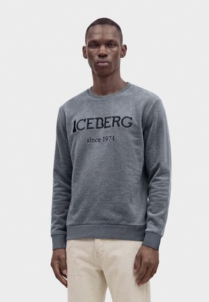 Млад мъж, облечен в сив суитшърт с дълги ръкави с текст "ICEBERG since 1974", комбиниран със светлобежови панталони на плътен фон.