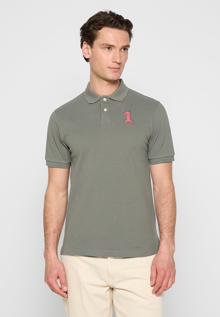 Man with light skin and dark hair wearing a gray-green polo shirt with a red "1" logo on the chest and beige pants, standing against a plain background.