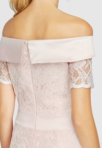 Vaaleanpunainen pitsimekko, jossa on olkaimeton satiinikaulus ja lyhyet pitsihihat, joissa on herkkiä kukkakuvioita ja vartalonmyötäinen siluetti.