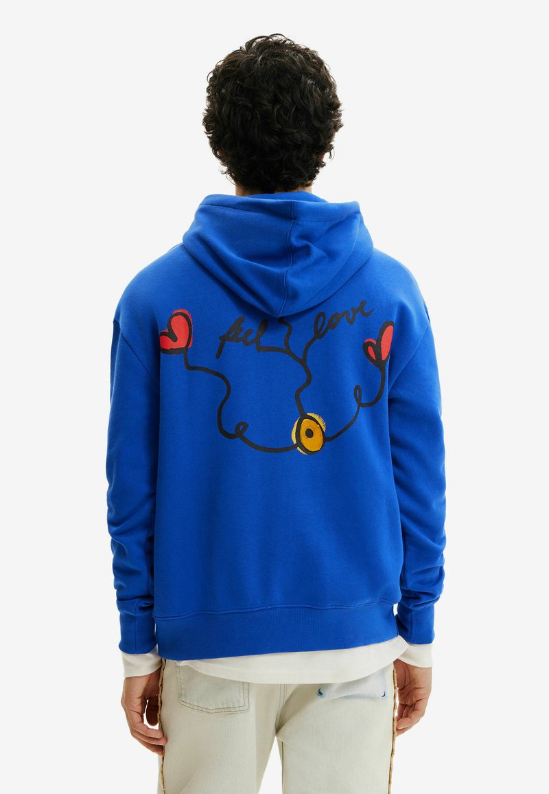 Mężczyzna w jasnoniebieskiej bluzie z kapturem z napisem "feel love", czerwonymi sercami i żółtym okręgiem na plecach, w połączeniu z jasnobeżowymi spodniami.