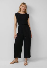 Schwarzer plissierter Jumpsuit mit rundem Halsausschnitt, kurzen Ärmeln und elastischem Taillenband. Kombiniert mit flachen, sandalenartigen Schuhen.