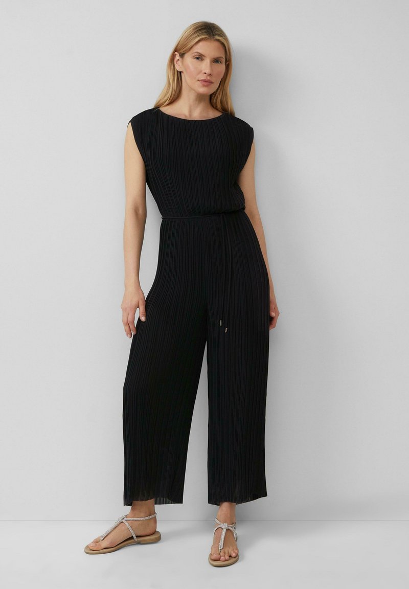 Schwarzer plissierter Jumpsuit mit rundem Halsausschnitt, kurzen Ärmeln und elastischem Taillenband. Kombiniert mit flachen, sandalenartigen Schuhen.