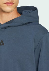 Hoodie aus dunkelblauem Stoff mit glatter Textur. Hat ein großes, schwarzes Logo auf der Vorderseite. Nahaufnahme des Ausschnitts.