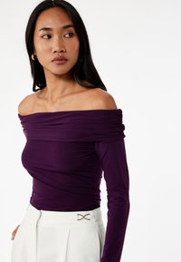 Väri violetissa off-the-shoulder -pusero pehmeästä ja joustavasta kankaasta, jossa on keräilyyksityiskohtia ja pitkät hihat, yhdistetty korkeavyötäröisiin valkoisiin housuihin.