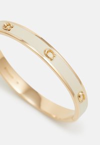 Bracciale in metallo dorato con un centro in smalto avorio liscio, caratterizzato da dettagli in rilievo a forma di bocca di cavallo, creando un design elegante e lucido.