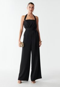 Woman wearing a sleeveless black halter jumpsuit with wide legs, holding a beige clutch and wearing gold sandals and jewelry.