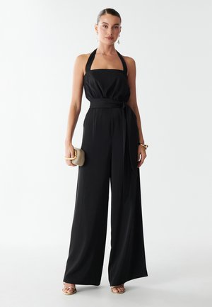 Kvinde iført en ærmeløs sort halter jumpsuit med brede ben, holder en beige clutch og har gyldne sandaler og smykker på.