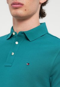 Πράσινη μπλούζα πόλο με υφασμάτινη υφή, με γιακά και τρία λευκά κουμπιά, διακοσμημένη με μικρό λογότυπο σε κόκκινο, λευκό και μπλε.