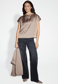 Top in satin a maniche corte color taupe, con scollatura ampia. Abbinato a jeans svasati scuri e in mano tiene una giacca coordinata.