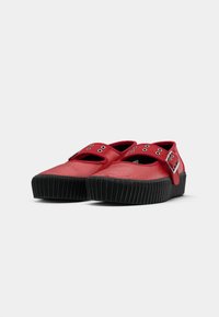 Par rde slip-on čevlje z debelimi črnimi rebrastimi podplati in srebrnimi zaponkami na belem ozadju.